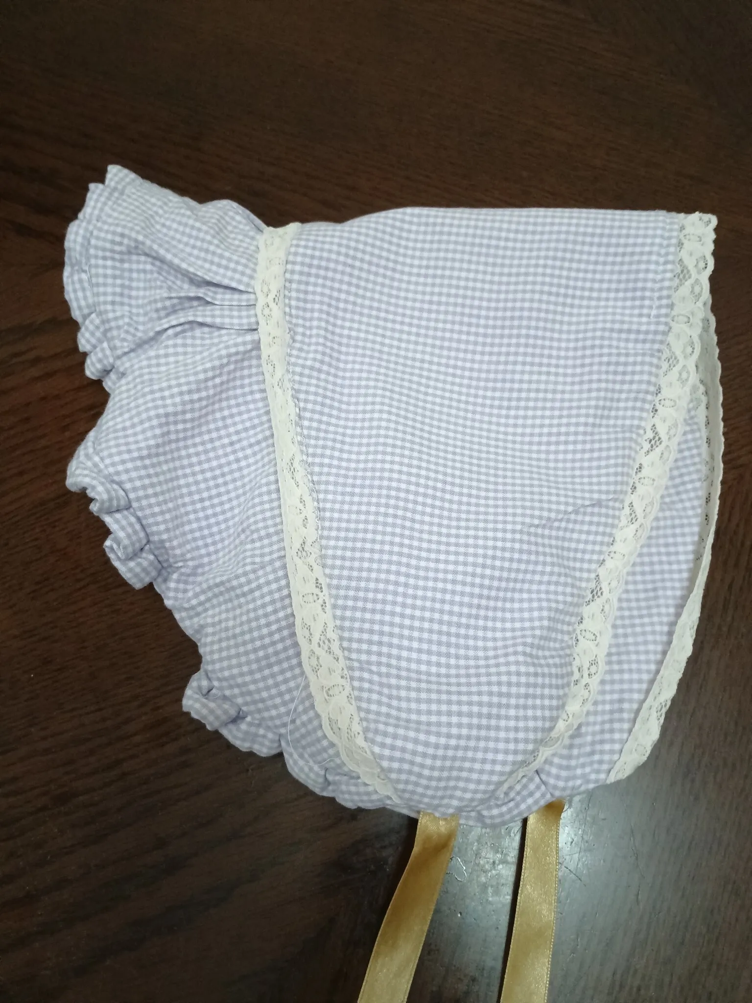 The bonnet lying flat on a wooden table: lavender gingham, gathered at the back, with cream lace along the brim and two gold satin ribbons.